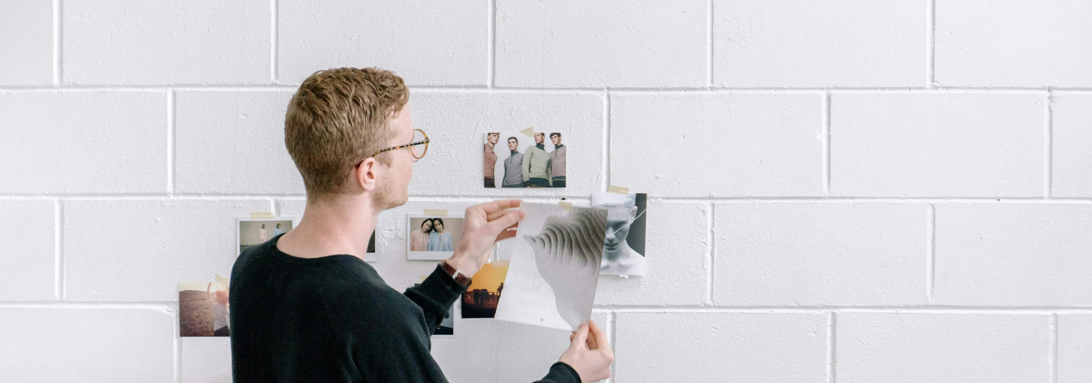 A man carefully arranges photographs on a white brick wall, thoughtfully curating the display. His focused expression and precise placement suggest he’s working on a creative project.