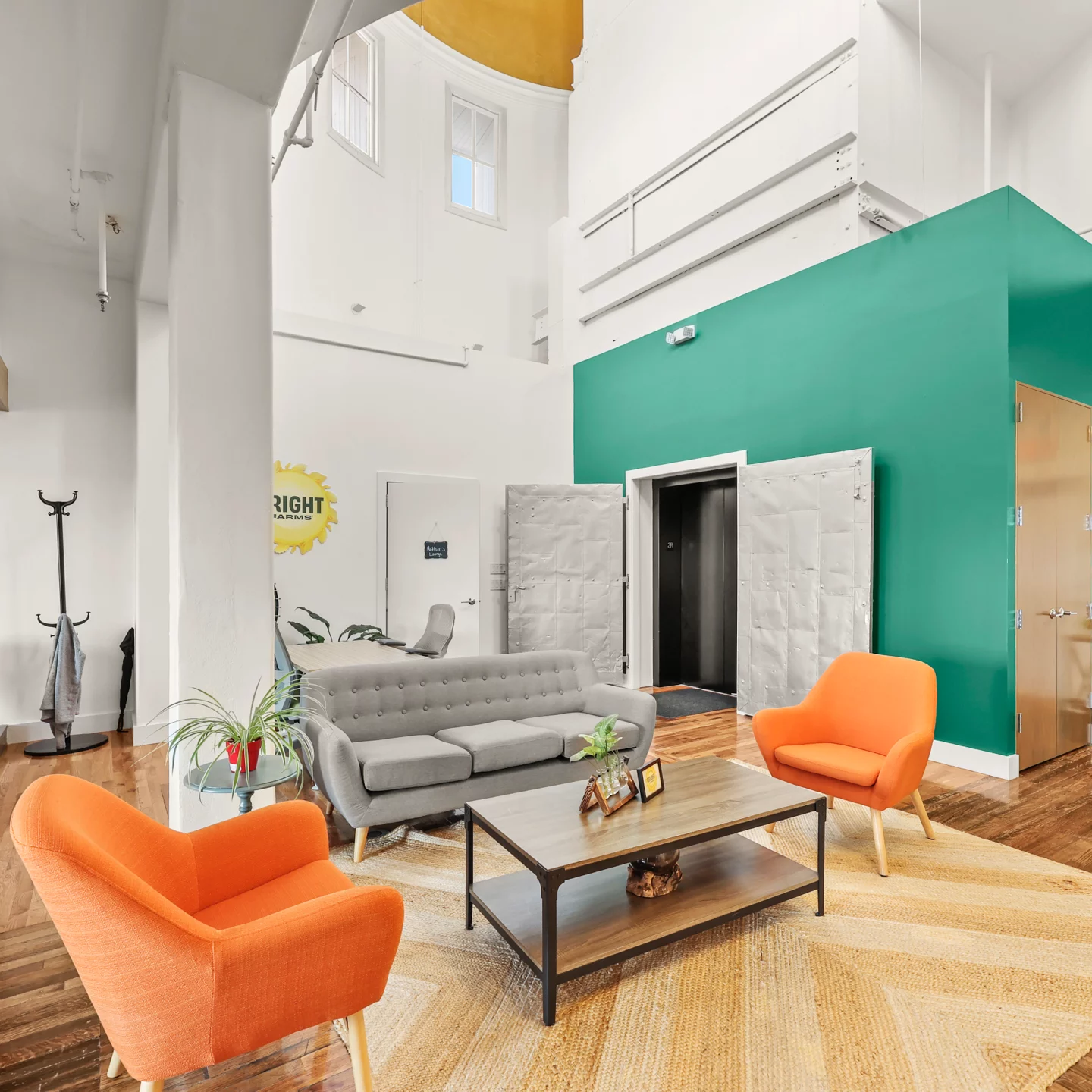 A modern coworking lounge with high ceilings, a green accent wall, and a mix of contemporary and mid-century furniture, including a gray sofa, two orange armchairs, and a wooden coffee table, creating a warm and inviting atmosphere.