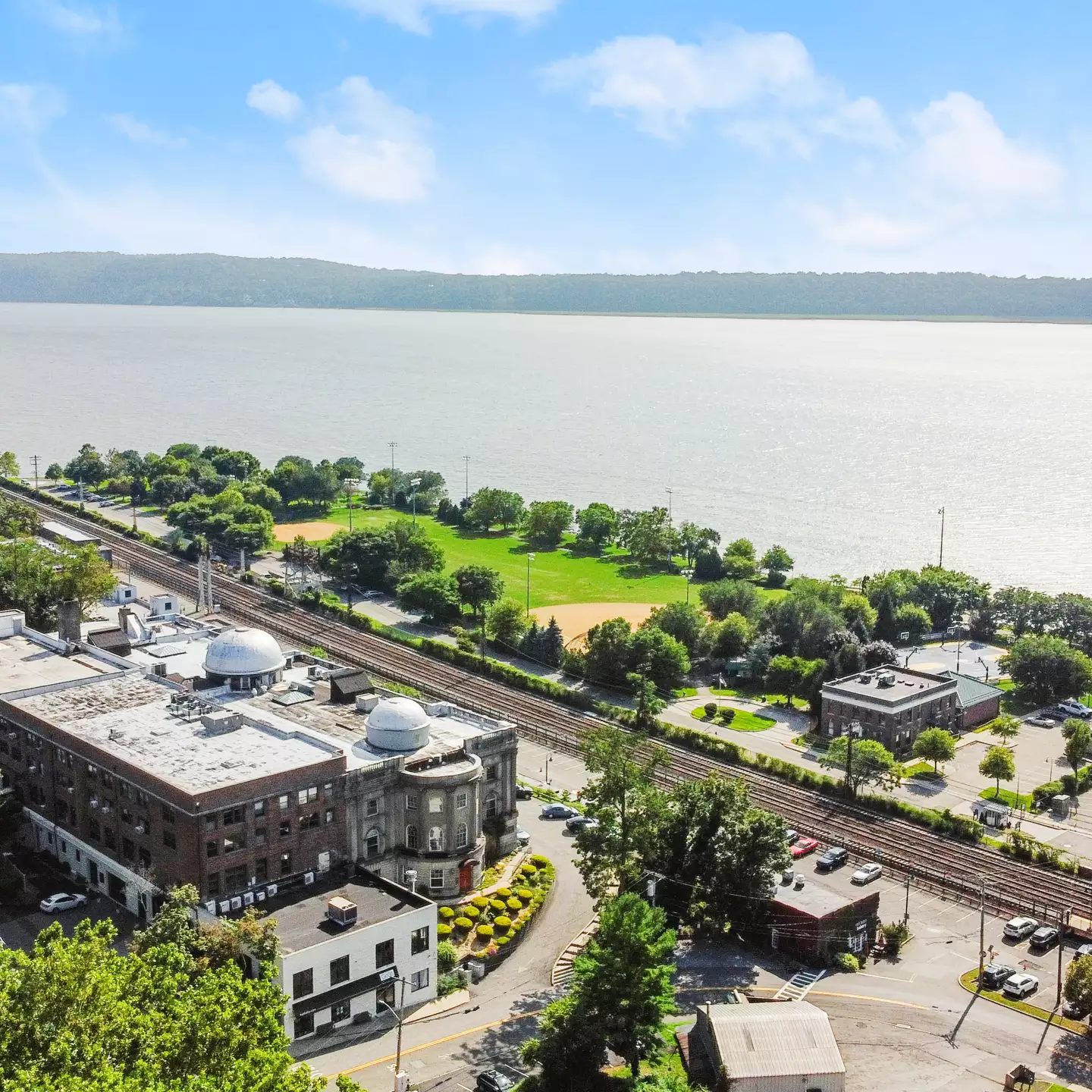 Aerial view of Irvington, NY, featuring a historic brick building near the Metro-North train tracks, with the Hudson River and lush green parkland stretching into the horizon under a bright blue sky.