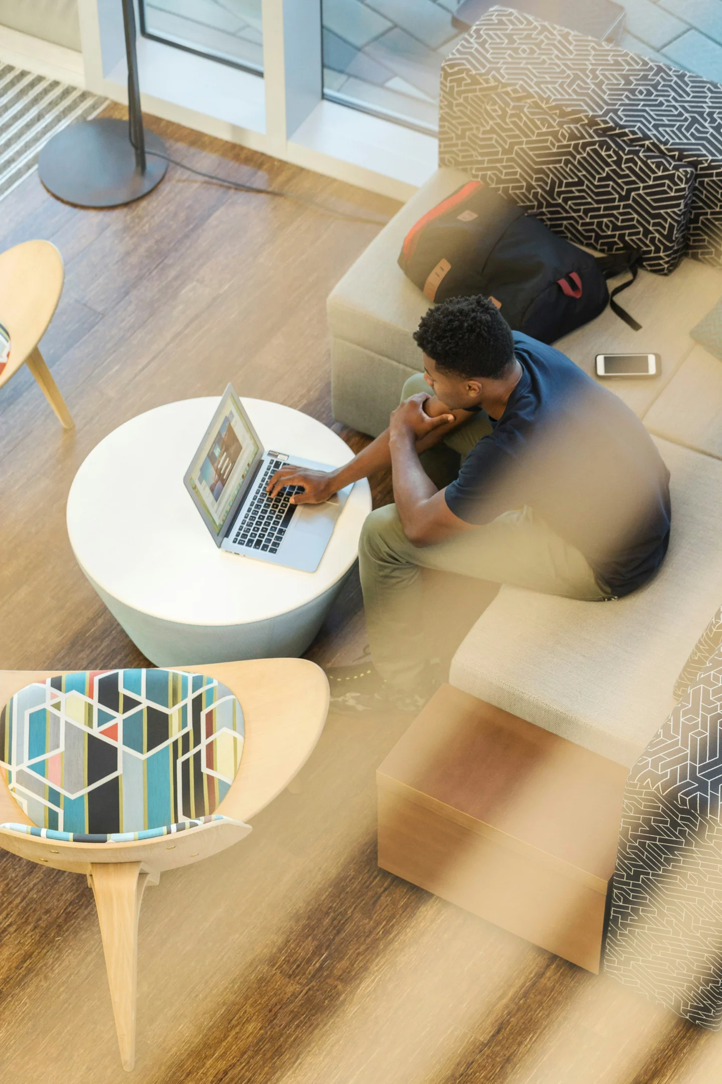 A young professional sits comfortably on a sofa, working on his laptop placed on a round table. Natural light filters through nearby windows, creating a relaxed yet productive atmosphere.