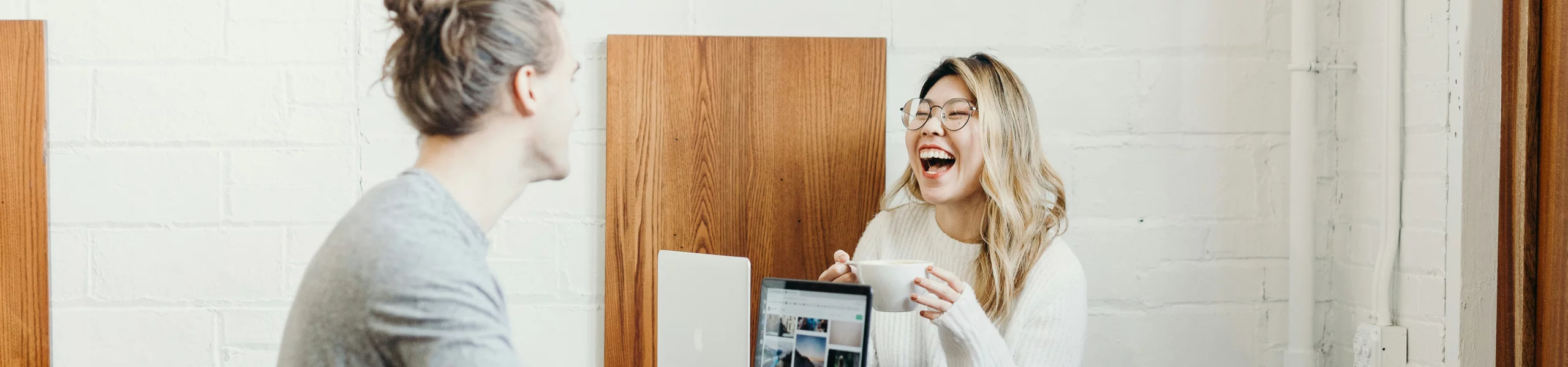 A man and a woman are engaged in a conversation, with the woman laughing while holding a cup in her hand. They appear to be enjoying a pleasant break together, sharing a lighthearted moment. The setting suggests a casual, friendly workspace atmosphere.