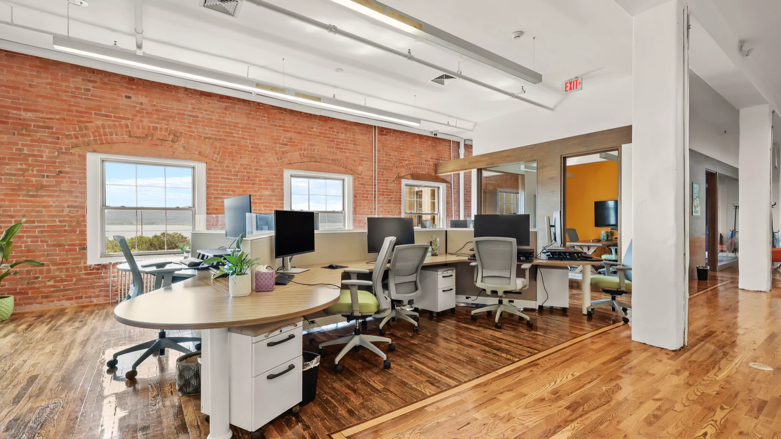 A professional and clean office setup featuring a table with desktop computers and chairs. The workspace faces a brick wall with large windows, offering scenic Hudson River views and an abundance of natural light.