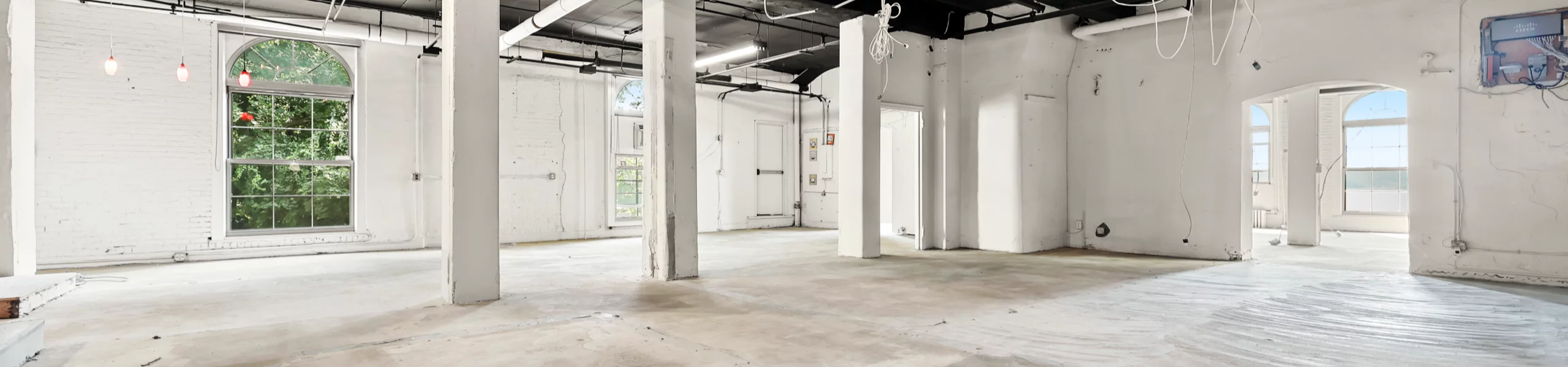 A bright, open workspace with expansive windows and high ceilings that invite natural light. The room’s minimalist state highlights its generous space and adaptable layout, offering a glimpse into its evolving transformation.